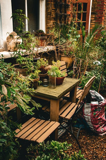 A small, cozy outdoor garden space adjacent to a brick house with a large window. The area features a rectangular wooden garden table surrounded by four matching wooden and metal chairs, with one part